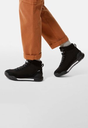 Zapatillas negras de caña alta con tejido de malla y acentos de goma, que cuentan con suelas texturizadas y cordones negros, combinadas con pantalones naranjas de puños enrollados.