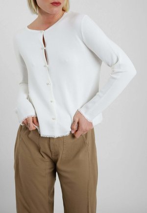 Persona che indossa un top bianco a maniche lunghe con bottoni perlati e bordi sfrangiati, abbinato a pantaloni marroni larghi, su sfondo chiaro.