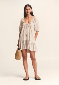 Abito a righe nei toni beige e crema con scollatura arricciata, maniche a puff corte e orlo arricciato. Una borsa di paglia e sandali piatti completano il look.