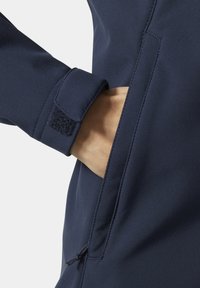 Ręka wewnątrz bocznej kieszeni granatowej kurtki z zapięciem na rzep przy mankiecie i widocznymi detalami szwów.