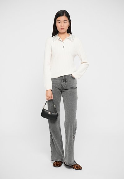 Kvinde står iført en hvid strikket sweater med udsmykket krave, grå jeans med sideskæringer, leopardmønstrede sko og holder en lille sort håndtaske.