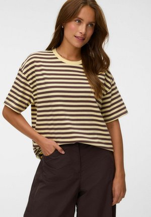 Femme portant une chemise à manches courtes rayée jaune et marron et un pantalon marron foncé, une main dans la poche, souriant légèrement.