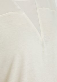 Světle béžový top s texturovanou látkou a průhledným překryvem, s V-výstřihem. Hladký povrch s jemným leskem.