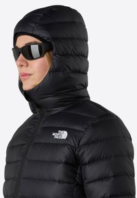 Svart puffjacka med hög huva, med en dragkedja framtill och horisontella quiltade sektioner. Logotyp på vänster bröst.