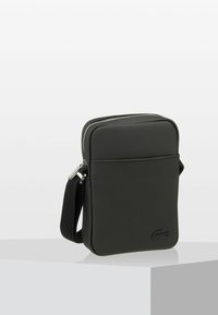 Black rectangular bag made of textured synthetic material. Features a top zipper, adjustable strap, and a front pocket. Lacoste logo on front.
