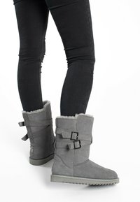 Botas de ante gris hasta el tobillo con forro de piel, que presentan dos correas con hebillas y suelas de goma texturizadas, combinadas con jeans ajustados negros.