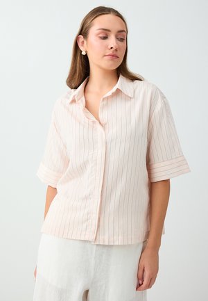 Jeune femme portant une chemise rose clair à manches courtes avec de fines rayures verticales et un pantalon blanc, debout devant un fond uni.