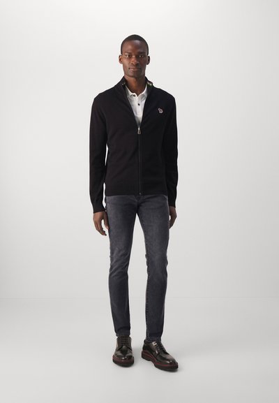 Black zip-up cardigan over a white polo, paired with dark jeans and black shoes. Cardigan features a colorful collar stripe and logo.