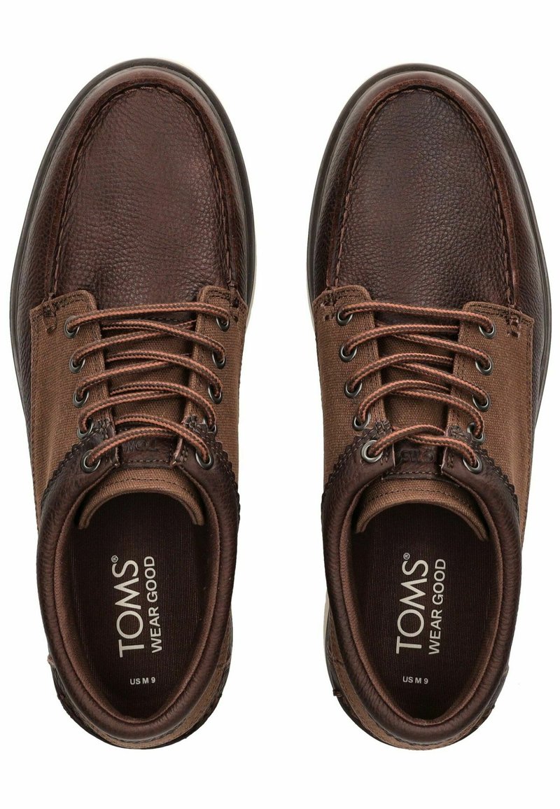 Zapatos de cuero y tela marrones con punteras redondeadas, cierre de cordones en la parte delantera y texturas contrastantes. Logo destacado en la plantilla interior.
