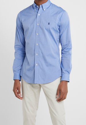 Mann trägt ein hellblaues, langärmliges Hemd mit Knöpfen und Kragen sowie beigefarbene Hose, steht vor einem einfachen Hintergrund.