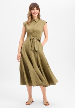 Femme souriante posant dans une robe midi sans manches vert olive boutonnée avec col, ceinture nouée à la taille et sandales plates beiges sur fond blanc.