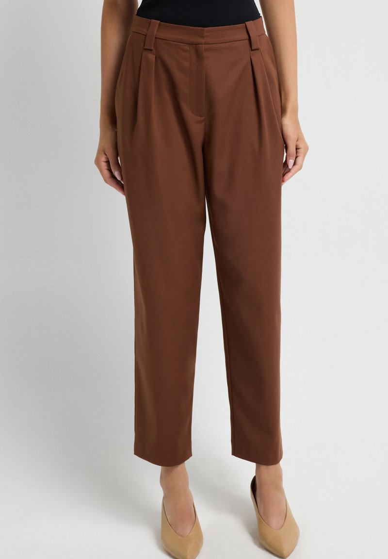 Vrouw in hoge, bruine, taps toelopende broek met plooien, gecombineerd met beige puntige hakken, staande tegen een effen witte achtergrond.