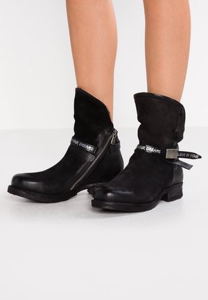 Czarne skórzane kozaki do połowy łydki z bocznymi zamkami i ozdobnymi paskami z napisem "WIERZ W SWOJE MARZENIA", noszone na gołych nogach.