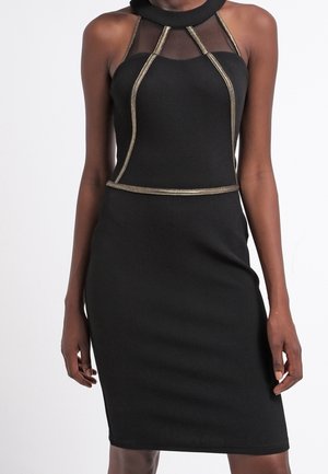 Czarna sukienka na ramiączkach, sięgająca do kolan, z przezroczystym dekoltem i złotą lamówką podkreślającą gorset i talię, noszona przez stojącą osobę.