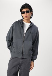 Grauer Reißverschluss-Hoodie aus weichem Material, mit einer vorderen Tasche, Kordelzugkapuze und abgenutztem Look. Kombiniert mit dunkelgrauen Hosen und Sonnenbrille.