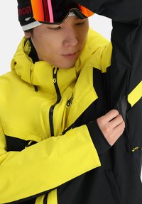 Veste de ski jaune et noire ; comprend une poche zippée, une capuche et un tissu texturé. Inclut un détail de logo et des poignets ajustables.