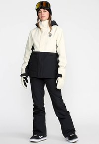 Completo da snowboard con giacca impermeabile crema e nera, pantaloni neri, guanti, occhiali da neve e scarpe da ginnastica nere.