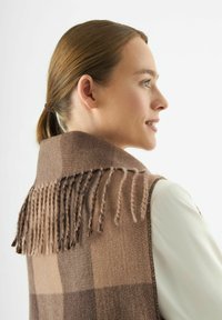 Gilet di lana marrone con dettaglio a frange lungo il colletto, caratterizzato da un motivo a quadri in tonalità più chiare. Abbinato a una camicia a maniche lunghe sotto.