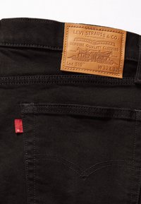 Los vaqueros de mezclilla negros presentan un parche trasero de cuero marrón con detalles de marca, una etiqueta roja en el lateral y costuras clásicas en los bolsillos.