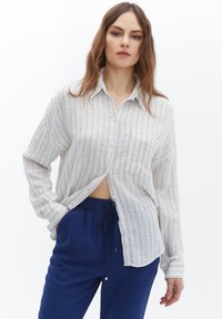 Lichtcreme knoopshirt met verticale blauwe strepen, lange mouwen en een relaxte pasvorm. Gepaarde met donkerblauwe, verstelbare broek.