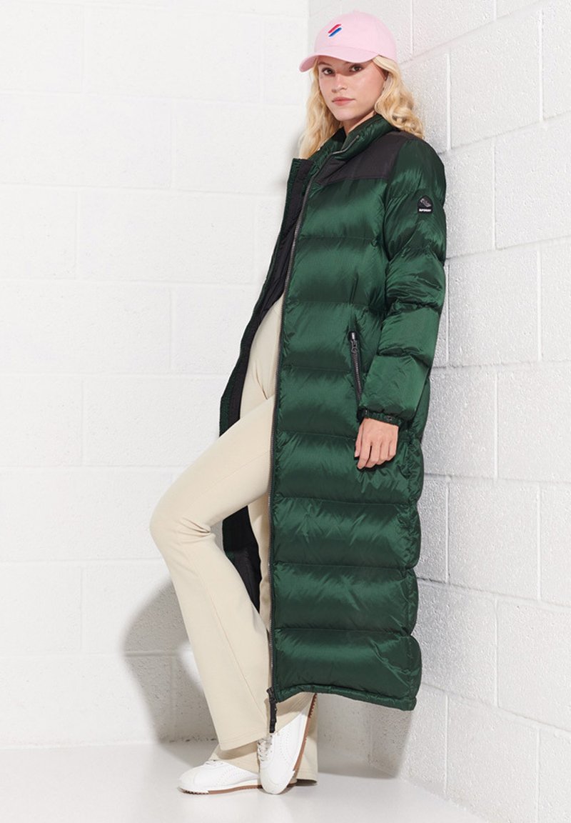Kobieta w długiej ciemnozielonej kurtce puchowej, beżowych spodniach, białych sneakersach i różowej czapce baseballowej, opierająca się o białą ceglany mur.