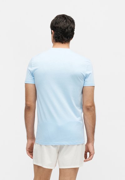 Άνδρας με σκούρα μαλλιά, φορώντας ανοιχτό μπλε κοντομάνικη μπλούζα και λευκά σορτς, στέκεται με την πλάτη προς ένα απλό λευκό φόντο.