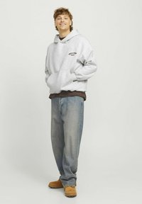 Sudadera gris con capucha y bolsillo delantero, sobre una camiseta marrón, combinada con jeans de color azul claro y zapatos marrones. Detalle de logotipo simple.