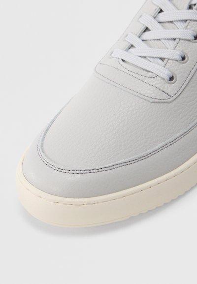 Αθλητικό παπούτσι από δέρμα ανοιχτού γκρι χρώματος με υφή στην επιφάνεια, στρογγυλεμένη μύτη και άσπρη λαστιχένια σόλα. Τα κορδόνια είναι ανοιχτού γκρι, με διακριτικές ραφές.