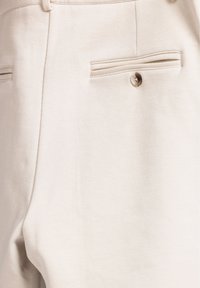 Beige op maat gemaakte broek met een zachte textuur, twee achterzakken met horizontale openingen en een klein knoopdetail op de rechterzak.