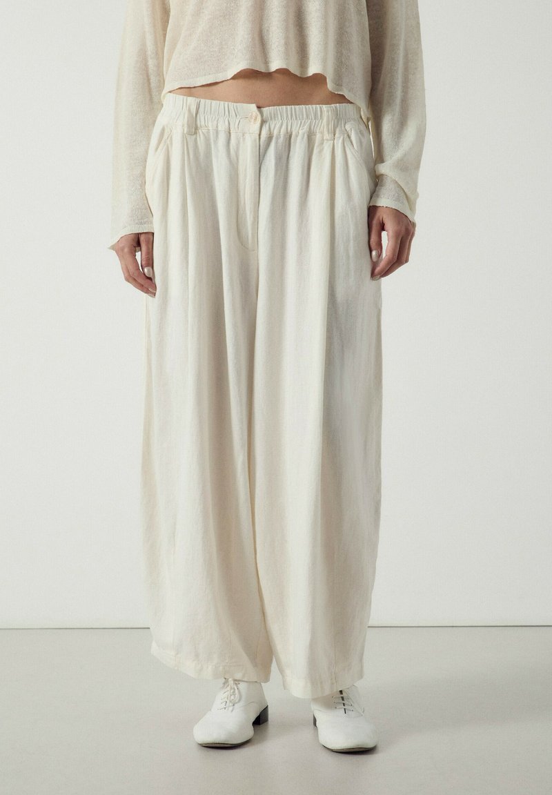 Femme portant un pantalon ample, taille haute, large et écru avec une ceinture froncée, associé à des chaussures blanches à lacets et un top court à manches longues.