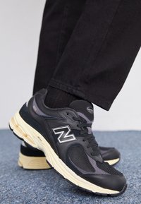 Chaussure de sport noire avec tige en mesh et cuir, arborant un logo "N" blanc, une semelle intermédiaire texturée et des lacets noirs. Chaussettes noires et pantalon sombre visibles.