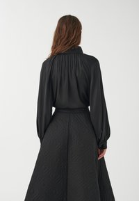 Blusa negra con espalda plisada y mangas largas, combinada con una falda negra acolchada que presenta un patrón geométrico. Textura suave.