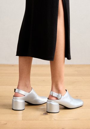 Ben iført en sort nederdel med sideslids og sølvfarvede blokhælede slingback-sko på et lyst trægulv.