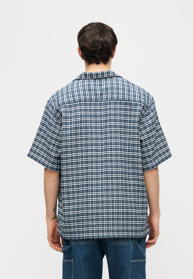 Persona vistiendo una camisa de manga corta a cuadros azul y blanca y jeans azules, vista desde atrás sobre un fondo claro y liso.