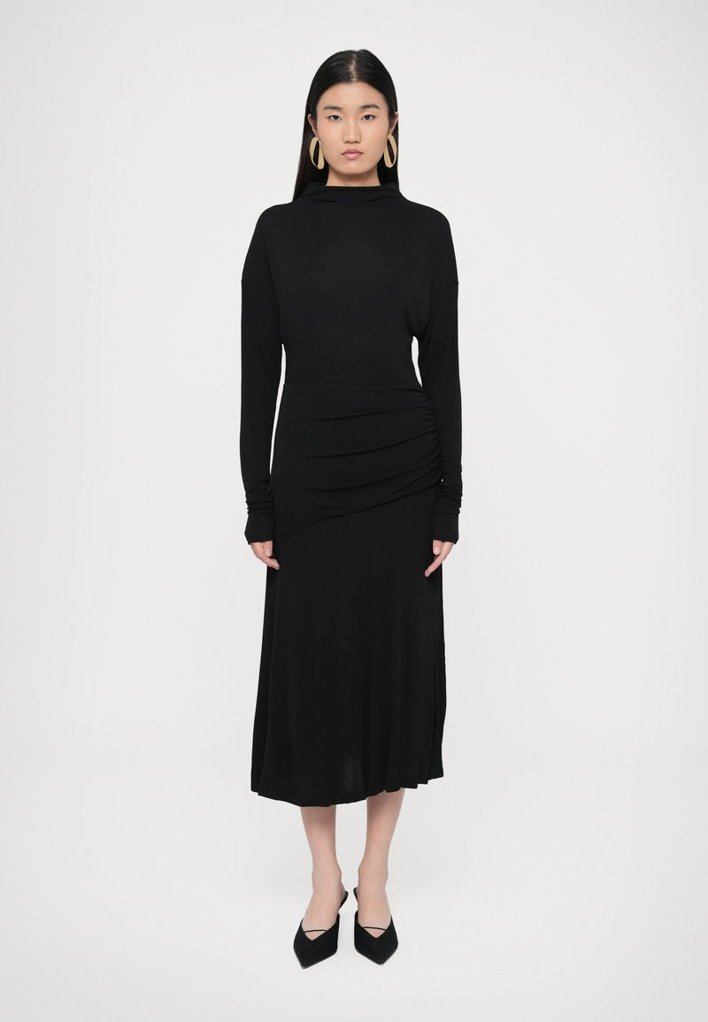 Fekete hosszú ujjú ruha, testhezálló fűzőrésszel, deréknál redőzött részlettel és bővülő szegéllyel. Feketére hegyesített cipővel és nagy fülbevalókkal párosítva.