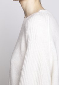 Biały sweter dziany o prążkowanej fakturze, wykonany z miękkiego materiału, o luźnym kroju, z okrągłym dekoltem i opuszczonymi ramionami.