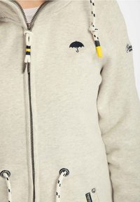 Hoodie zippé gris clair avec un logo de parapluie brodé, des cordons de contraste et des œillets renforcés pour plus de détails.