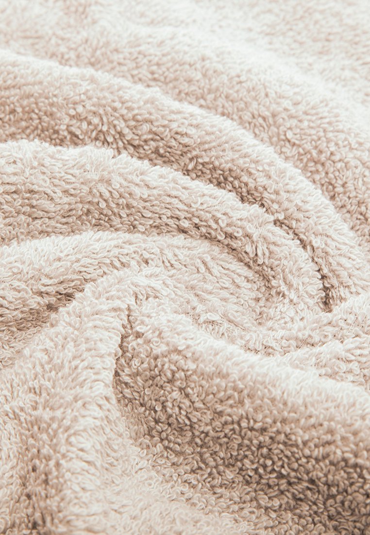 Beige Frottee-Handtuch mit einer weichen, strukturierten Oberfläche, das durch sein unregelmäßiges Erscheinungsbild die plüschigen Fasern und das geschlungenen Design hervorhebt.