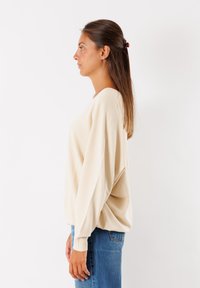 Maglione beige oversize con scollo ampio, tessuto testurizzato e polsini a costine, abbinato a jeans in denim blu. Modella mostrata in vista di profilo.