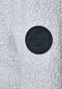 Grå ytterkläder med ett texturerat, fluffigt material. Har en svart cirkulär patch märkt "CECIL OUTERWEAR." Den släta texturen kontrasterar med det fluffiga tyget.