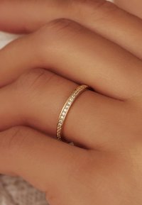 Goldring mit einem dünnen Band, das eine Reihe von kleinen klaren Kristallen und strukturierten Kanten zeigt, liegt auf einer Hand mit heller Haut.