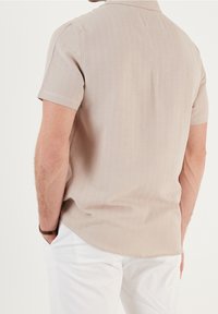 Camicia beige a maniche corte con colletto, texture liscia, orlo dritto e dettagli di tessitura fine. Indossata con pantaloni bianchi.