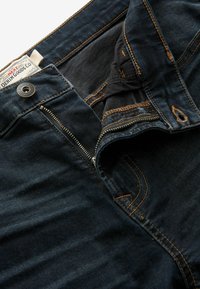 Tamno plavi traperice s zlatim šavovima, s patentnim zatvaračem, prednjim džepom i oznakom "DENIM GOODS CO." Teksturirana tkanina.