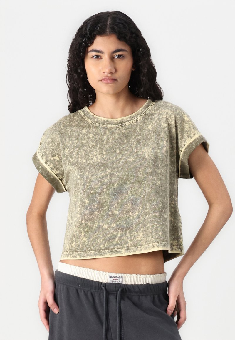 Kurzärmliges, cropped T-Shirt in einem gedämpften Olivgrün mit strukturiertem Finish und rohen Kanten. Kombiniert mit dunkelgrauen Hose mit elastischem Bund.