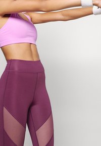 Fioletowe legginsy z wysokim stanem z siateczkowymi panelami oraz dopasowanym różowym stanikiem sportowym. Gładki materiał o eleganckim wzornictwie. Nosi się białe opaski na nadgarstki.