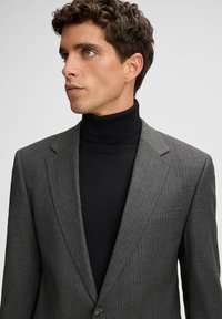 Junger Mann mit kurzen lockigen Haaren, der ein graues Nadelstreifen-Blazer über einem schwarzen Rollkragenpullover trägt, und seitlich gegen einen schlichten Hintergrund blickt.