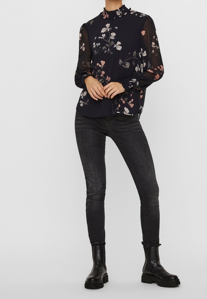 Femme portant un chemisier noir à fleurs avec col montant et manches transparentes, jean skinny foncé et bottines noires, debout les mains jointes.