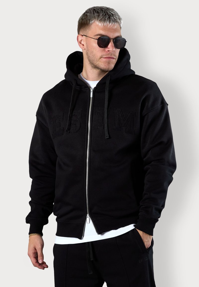 Schwarze Zip-Up-Hoodie mit strukturiertem Frontlogo, Kapuze mit Kordelzug, gerippten Bündchen und sichtbarem kontrastierendem weißen Unterhemd am unteren Rand.