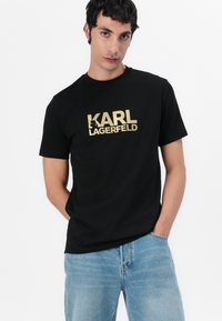 Camiseta de algodón negra con el logotipo "KARL LAGERFELD" impreso en dorado. Manga corta, cuello redondo, ajuste holgado. Combinada con pantalones vaqueros de mezclilla azul claro.