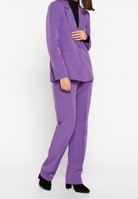 Lila skräddarsydd kostym med en enfaldig kavaj och byxor med raka ben, tillverkad av slät tyg med subtil glans. Svart polotröja under.